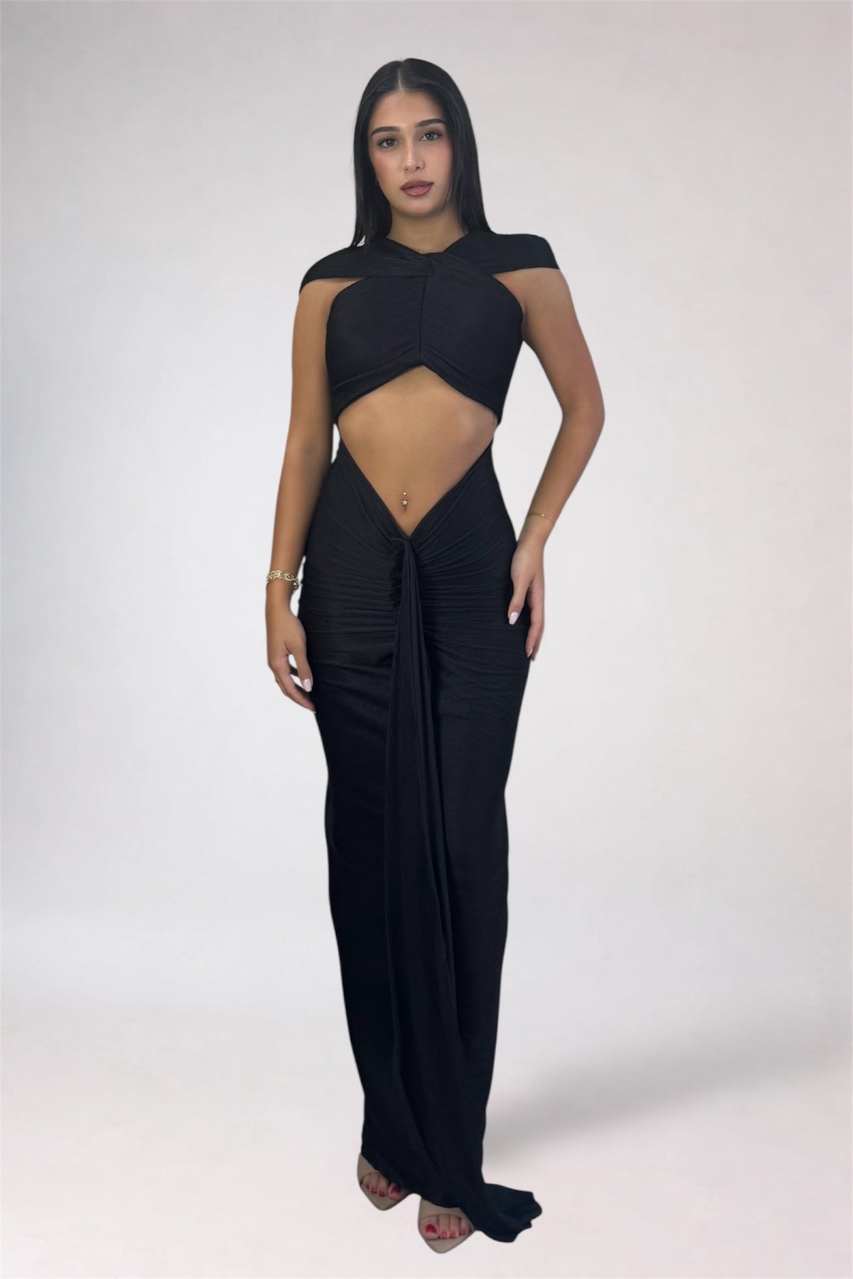 A woman modeling a black sleeveless maxi dress with a dramatic cutout at the waist and a draped skirt, photographed against a plain light gray studio background.