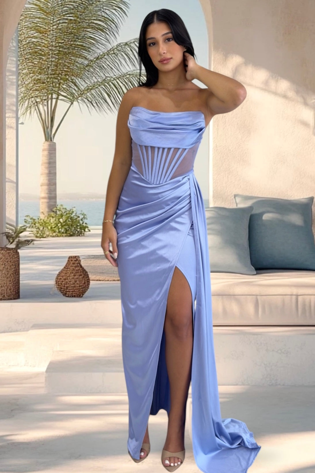 A woman models a light blue strapless maxi gown with a sheer corset-style bodice, draped skirt, and high front slit in an outdoor patio setting.