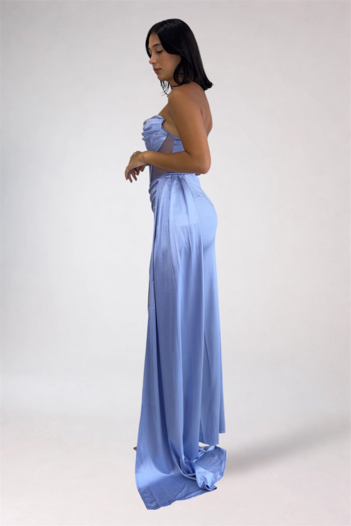 A woman modeling a strapless light blue satin maxi dress with a draped, ruffled bodice and flowing skirt, shown in profile against a plain light gray studio background.