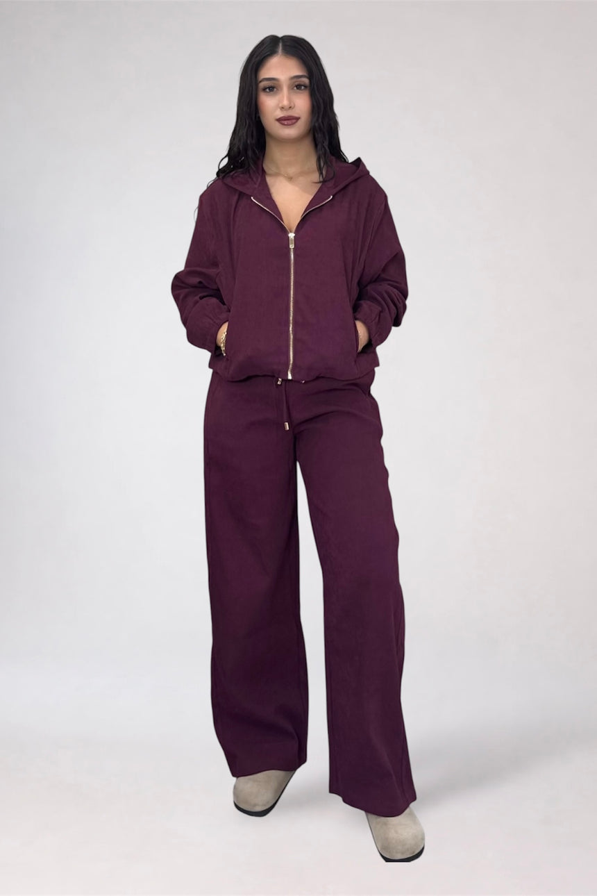 A woman modeling a matching deep plum purple tracksuit with a zip-up hoodie and wide-leg pants, standing against a light gray studio background.