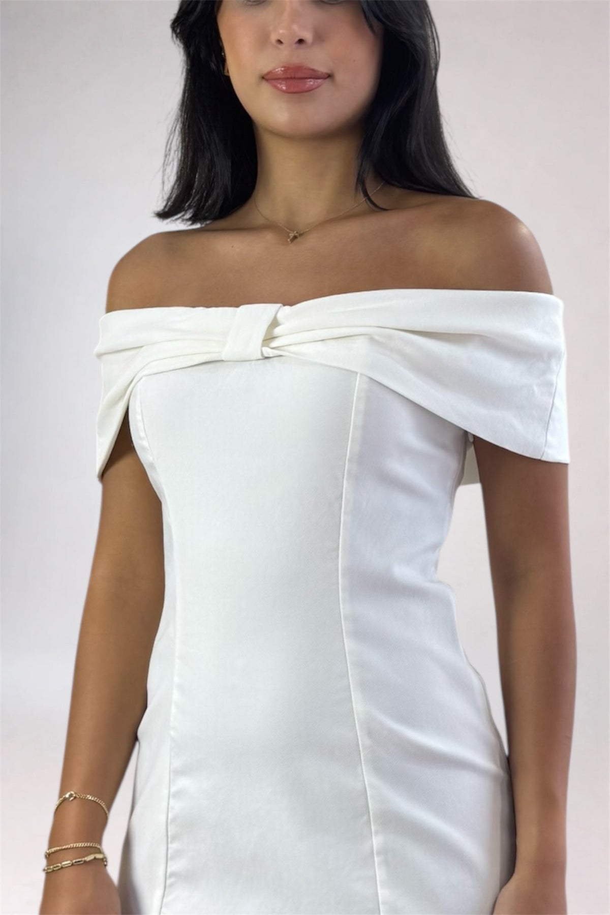 A woman wearing an off-the-shoulder white dress with a structured bodice and a large bow detail at the neckline, photographed against a plain light background.