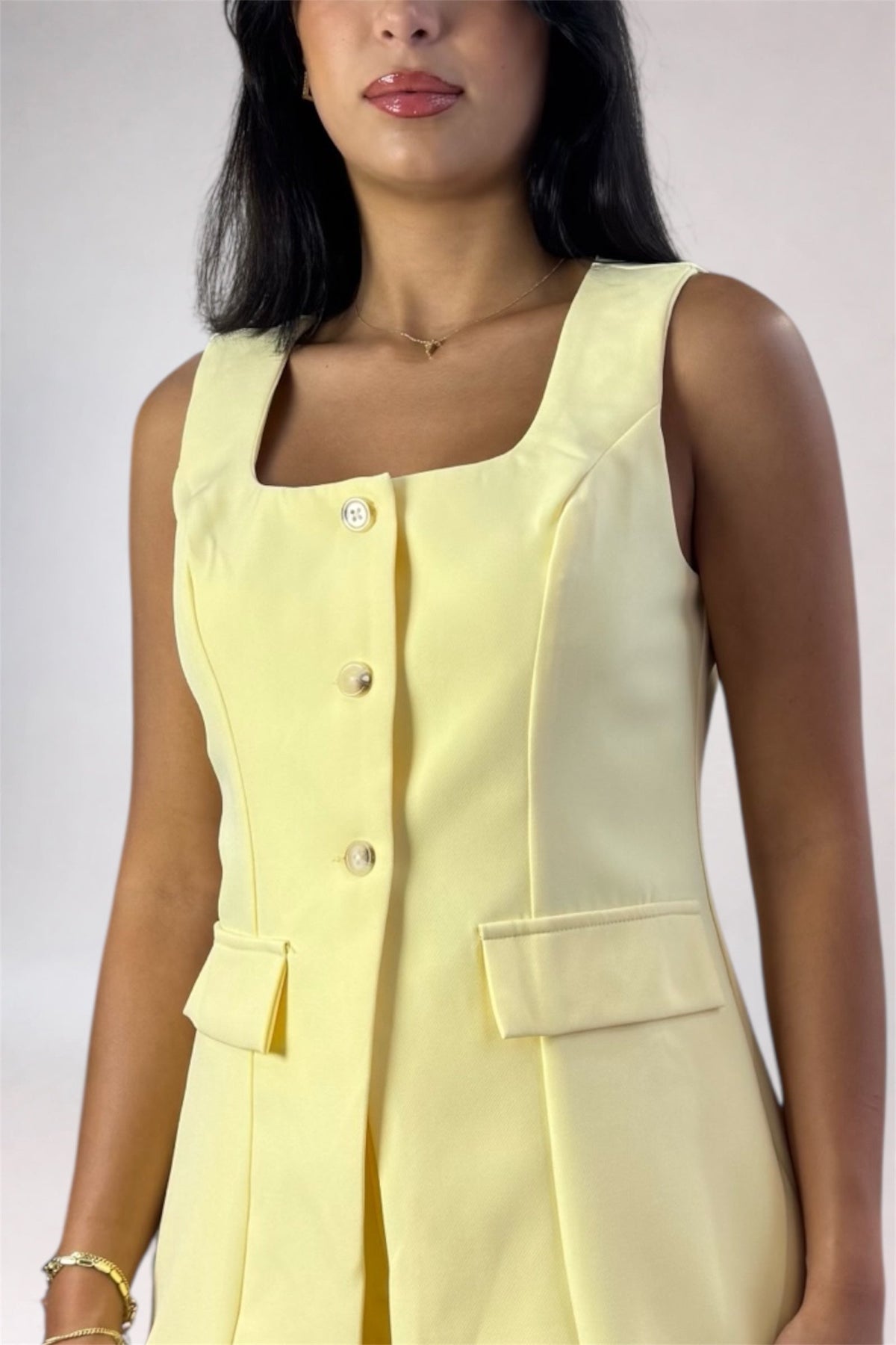 A woman wearing a pale yellow sleeveless dress with a square neckline, front button closure, and flap pockets, shown against a plain light background.