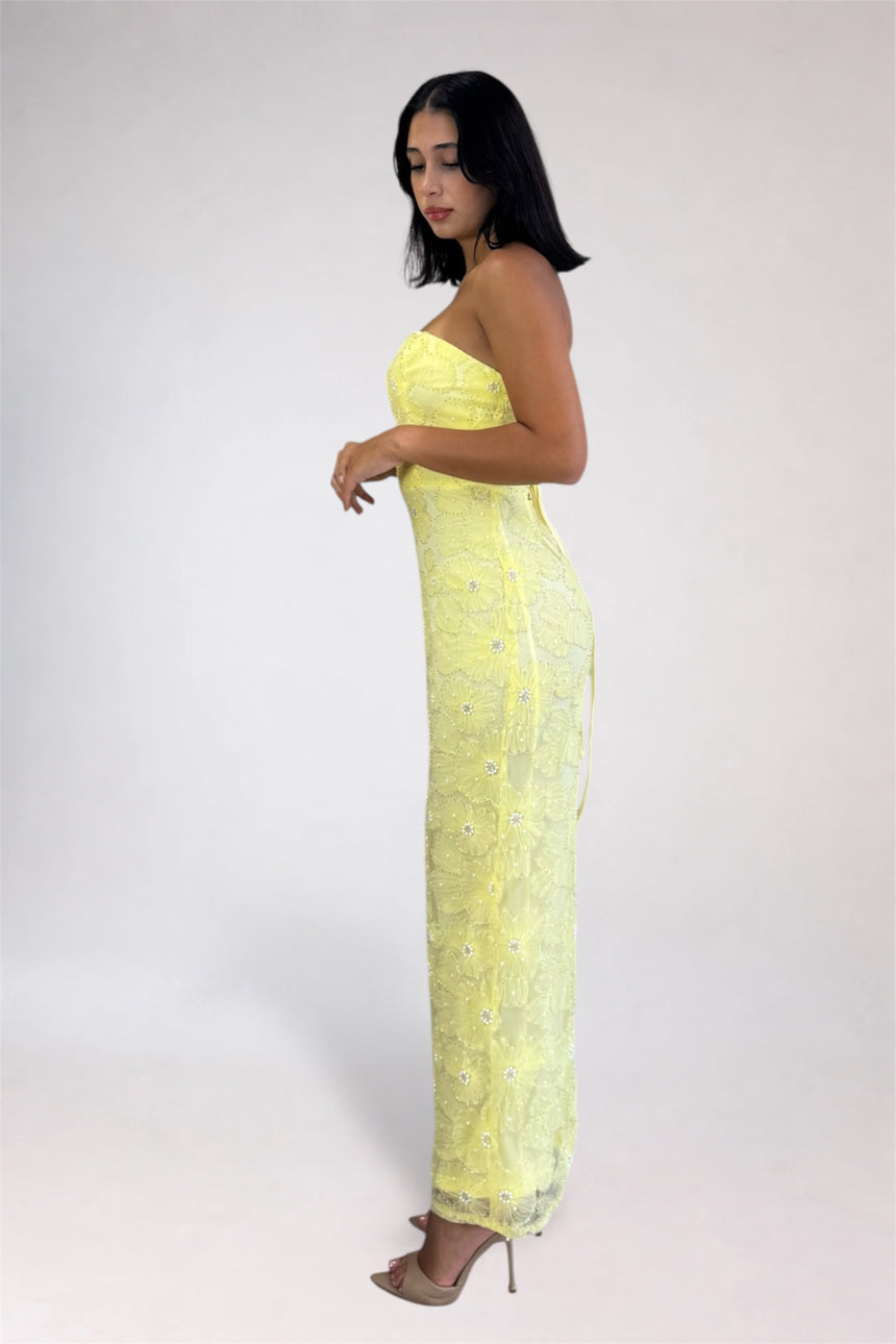 A woman modeling a strapless, floor-length yellow lace dress with silver embellishments, standing in profile against a light gray studio background.