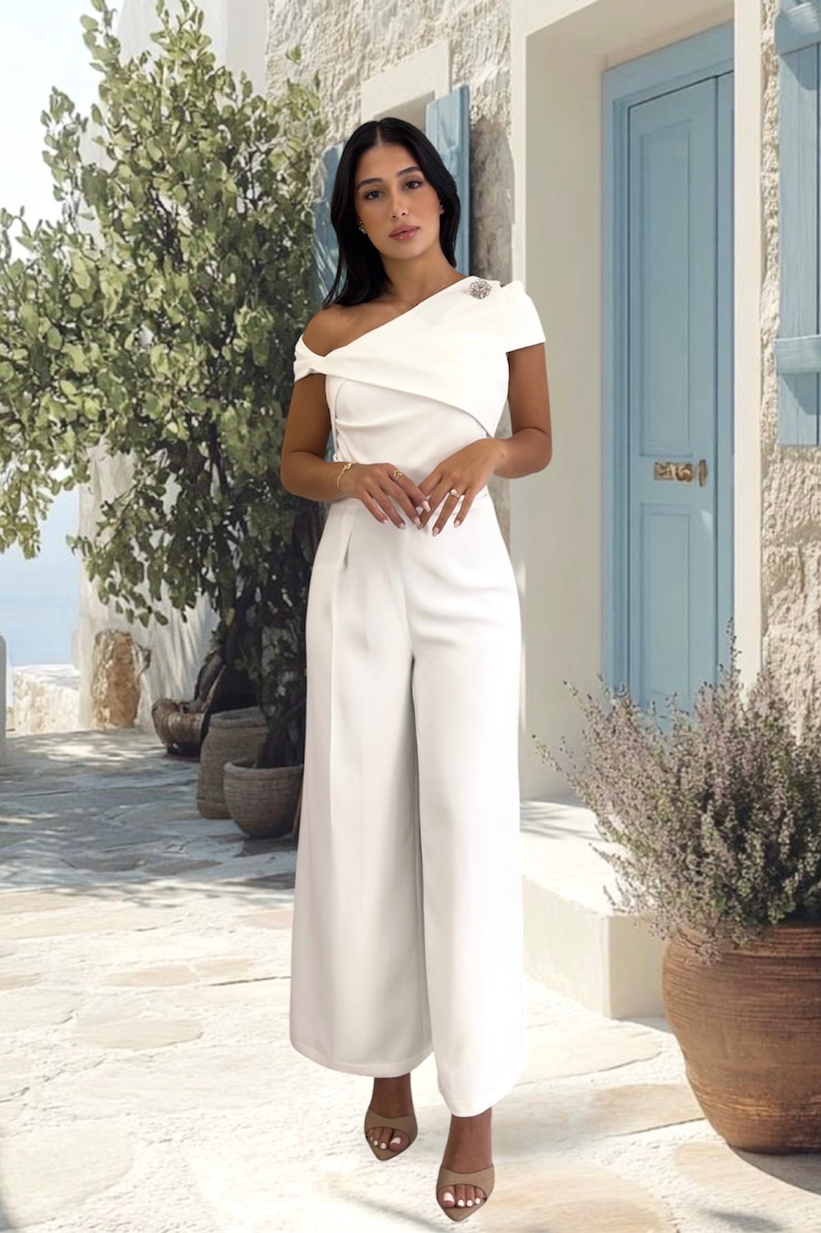 A woman modeling a white one-shoulder jumpsuit with wide-leg pants and nude high-heeled sandals against a light blue street