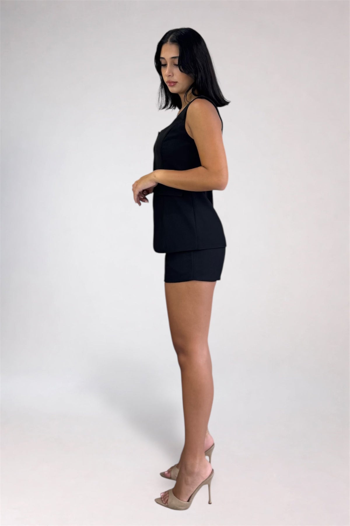 A woman modeling a black sleeveless top and matching black shorts set, paired with clear high-heeled sandals, against a plain light gray studio background.
