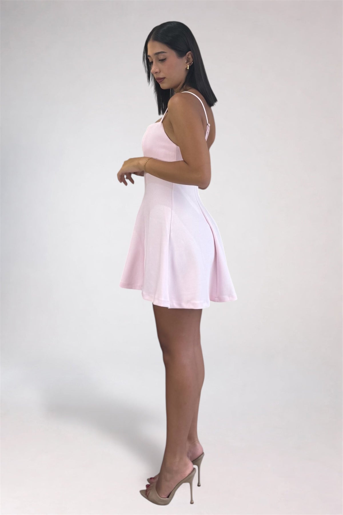A woman modeling a light pink sleeveless mini dress with thin straps and a flared skirt, paired with clear-strap high heels, against a plain light gray studio background.