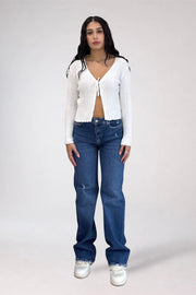 A model stands against a light gray studio background wearing a white ribbed long-sleeve tie-front cardigan, blue jeans, and white sneakers.