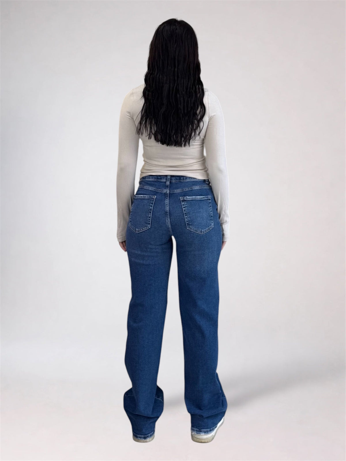 A person viewed from the back wearing a fitted light beige long-sleeve top and blue wide-leg jeans against a plain light gray studio background.