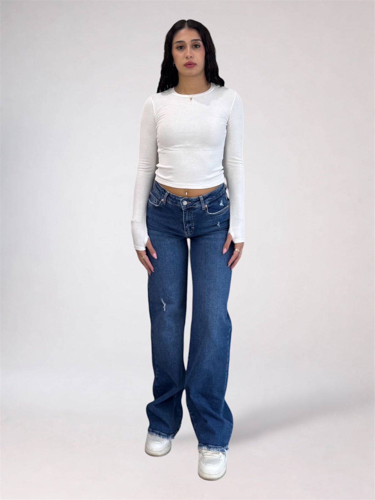 A model stands against a light gray studio background wearing a fitted white long-sleeve crop top, blue high-waisted straight-leg jeans with subtle distressing, and white sneakers.