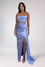A woman modeling a light blue satin strapless gown with a draped bodice, sheer mesh panels, a high thigh slit, and a long flowing train against a plain light gray studio background.