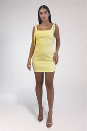 A woman modeling a pale yellow ribbed mini dress with square neckline and tie straps, paired with nude high-heeled sandals, against a light gray studio background.