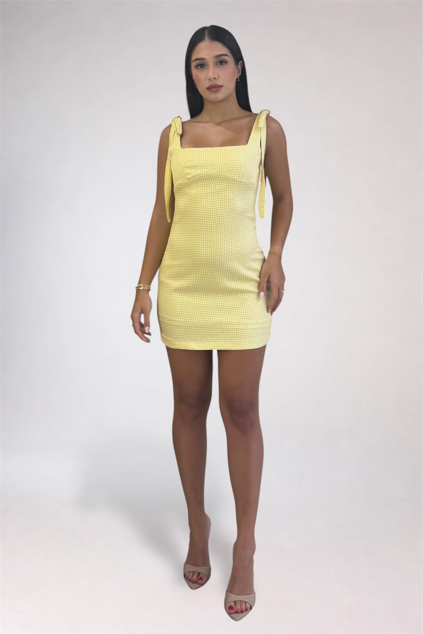 A woman modeling a pale yellow ribbed mini dress with square neckline and tie straps, paired with nude high-heeled sandals, against a light gray studio background.