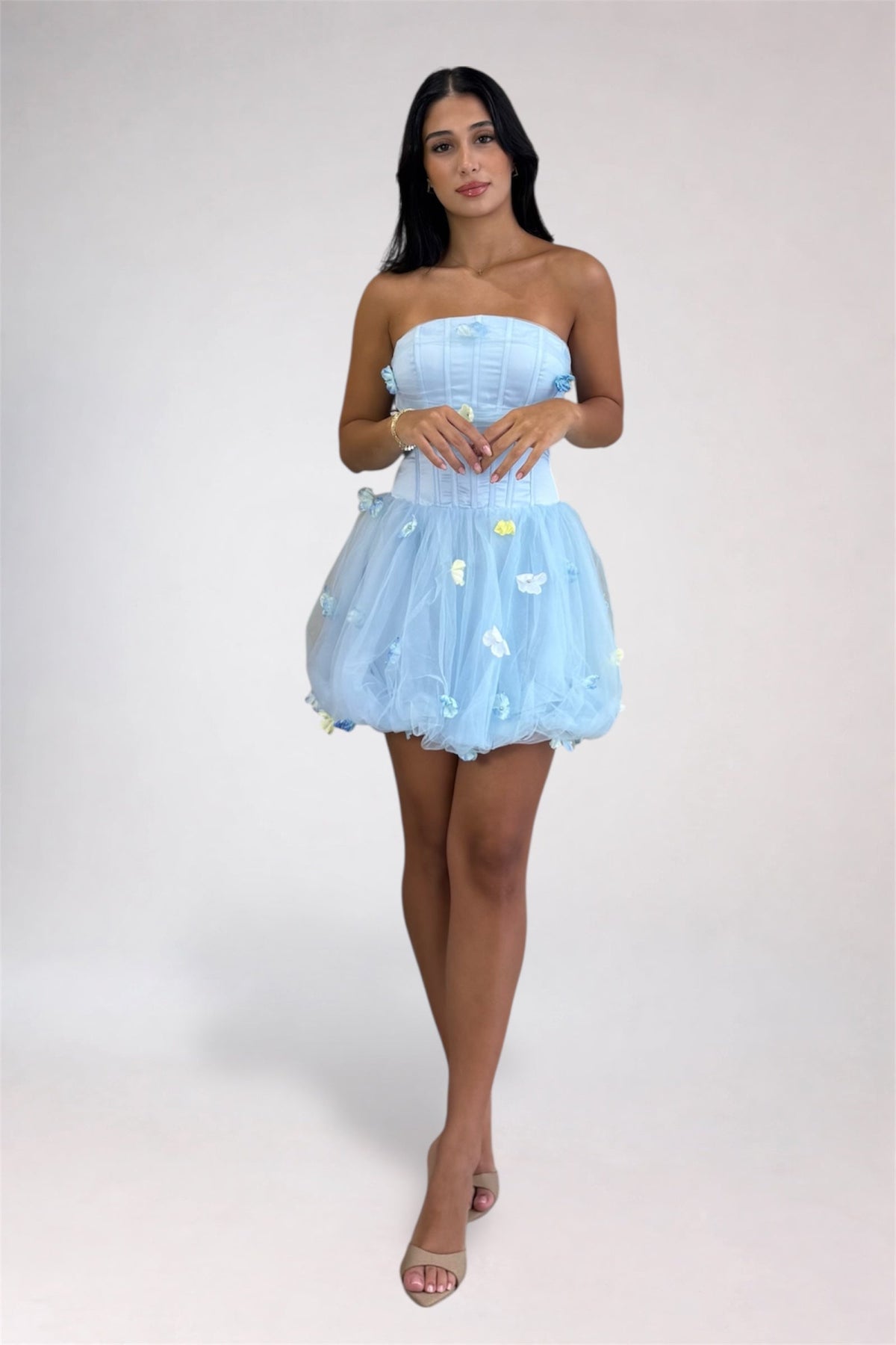 A woman modeling a strapless light blue tulle mini dress with a corset-style bodice and small white and yellow applique details, standing against a plain light gray studio background.