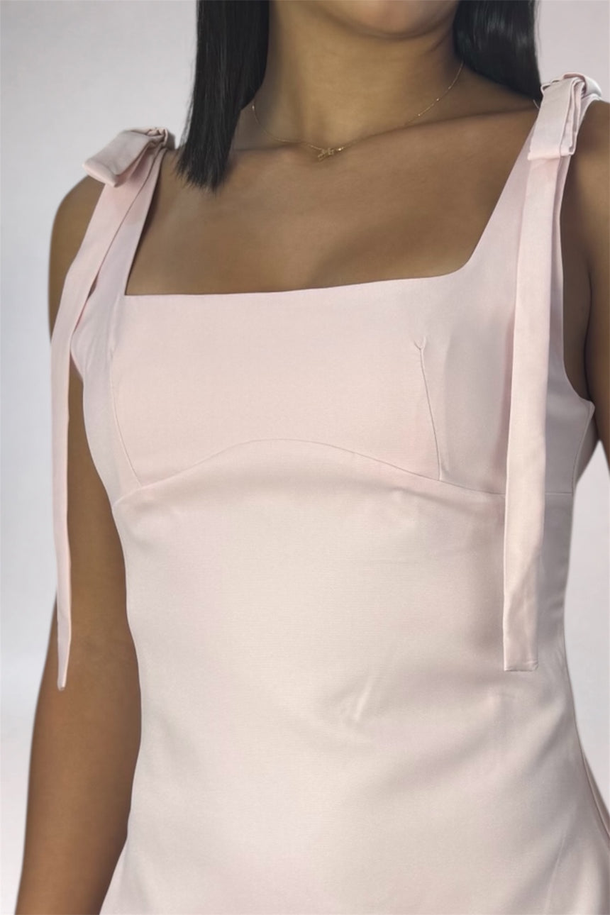 A close-up of a model wearing a pale pink satin-like dress with a square neckline, structured bodice, and thin shoulder ties with bow details.