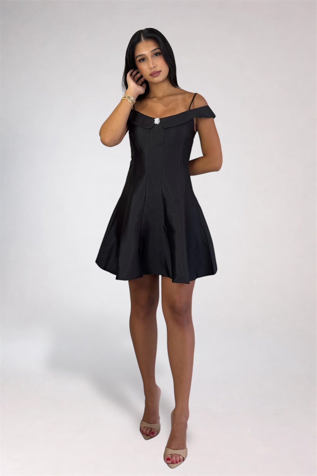A woman modeling a short black off-the-shoulder dress with a structured bodice and flared skirt, paired with nude high-heeled sandals, against a light gray studio background.