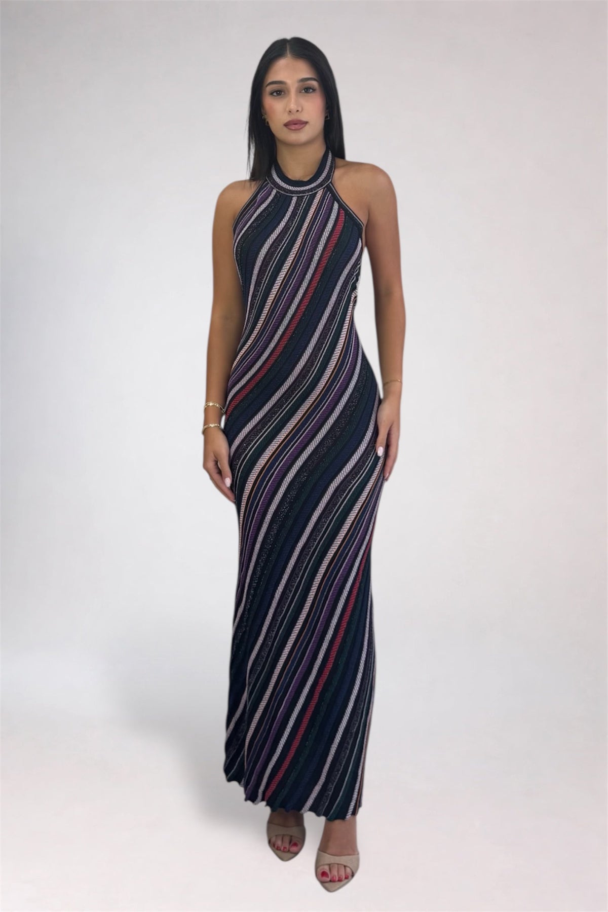 A woman modeling a sleeveless, high-neck maxi dress with diagonal stripes in navy blue, white, and red, standing against a light gray studio background.