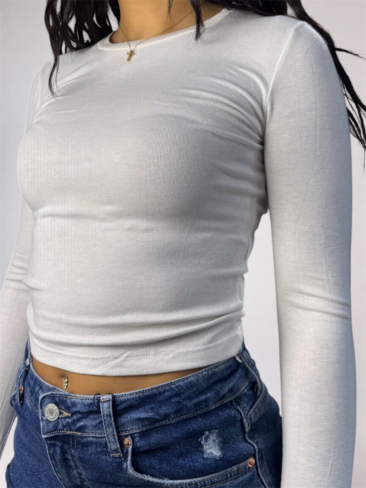 A woman wearing a fitted, long-sleeve white ribbed crop top and blue jeans, cropped from the neck down against a plain light background.