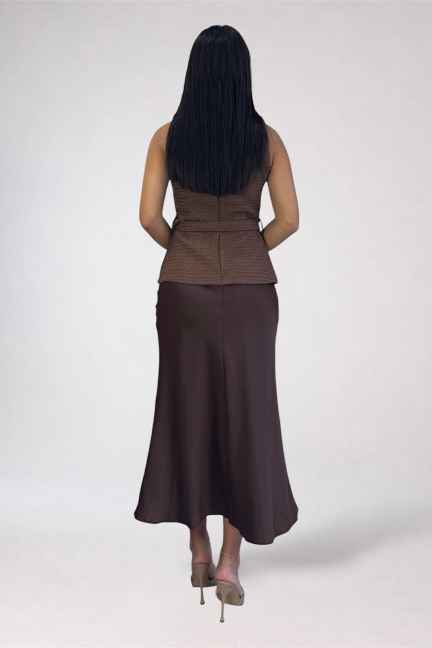A woman shown from the back wearing a sleeveless brown textured top and a long dark brown skirt, standing against a plain light gray studio background.