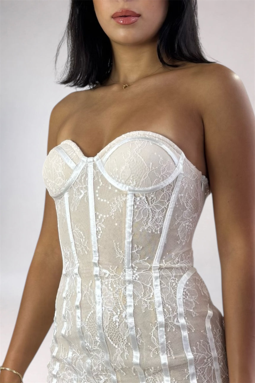 A close-up of a model wearing a strapless white lace corset with satin trim and underwire cups against a light gray studio background.