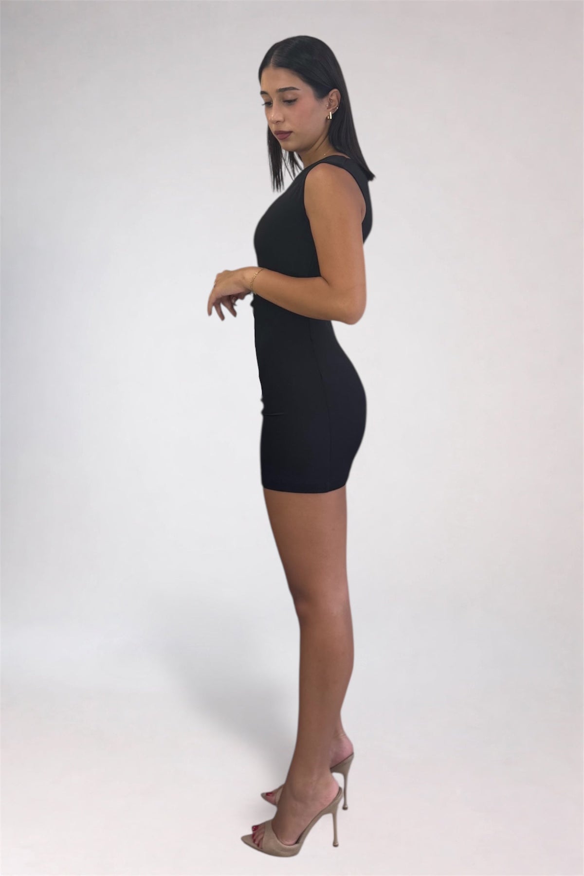 A woman modeling a sleeveless black bodycon mini dress with nude high-heeled sandals, standing in profile against a plain light gray studio background.