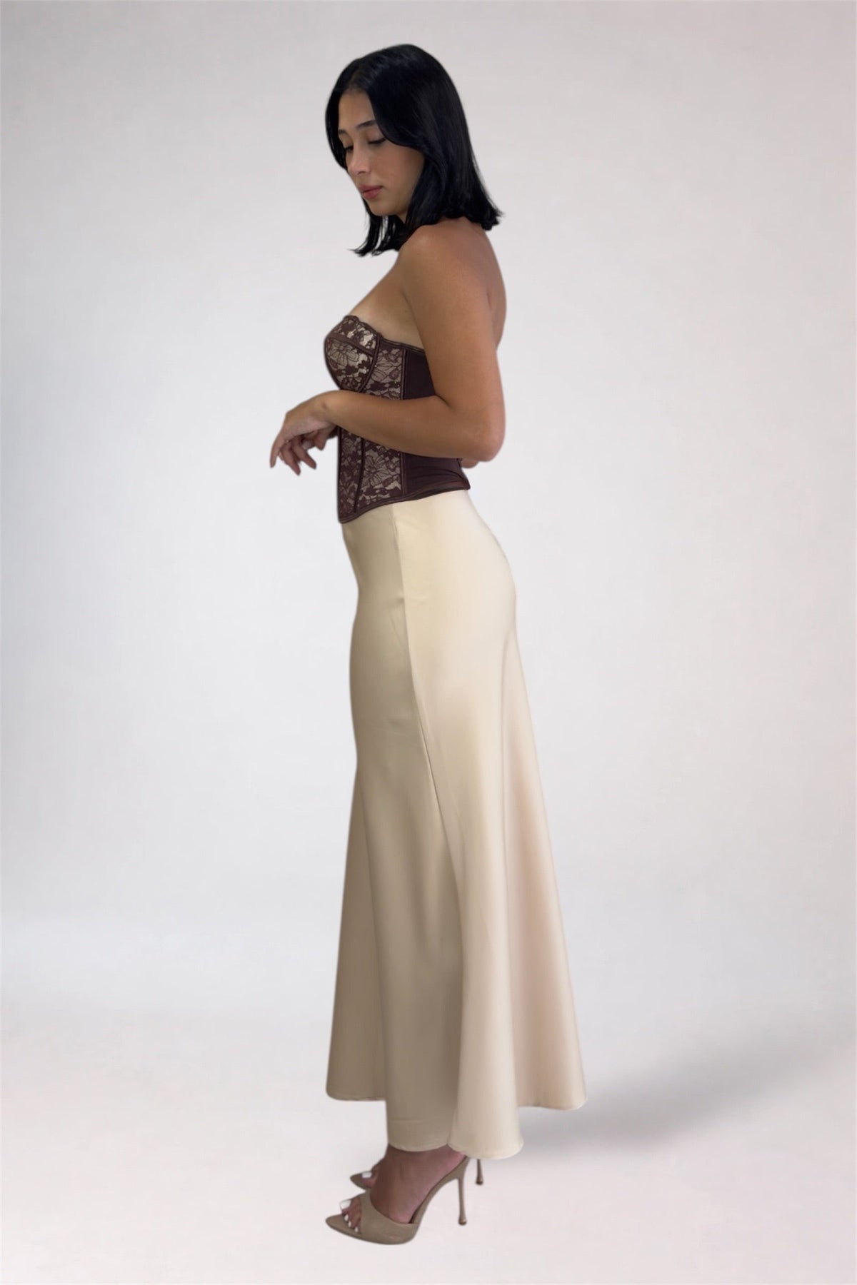 A woman modeling a strapless formal gown with a dark brown lace corset-style bodice and a flowing cream skirt, shown in profile against a plain light gray studio background.