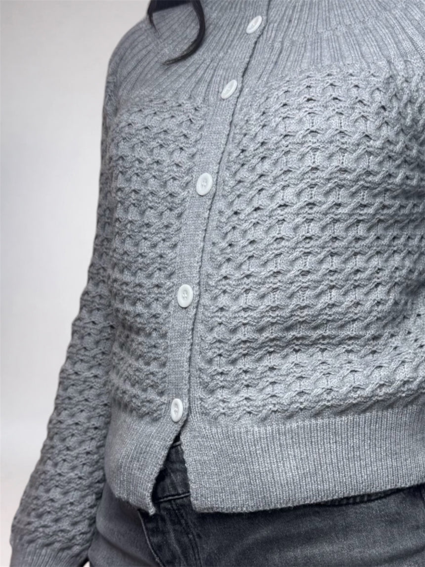 A close-up of a person wearing a gray textured knit cardigan with white buttons, paired with dark gray jeans, against a plain light gray background.
