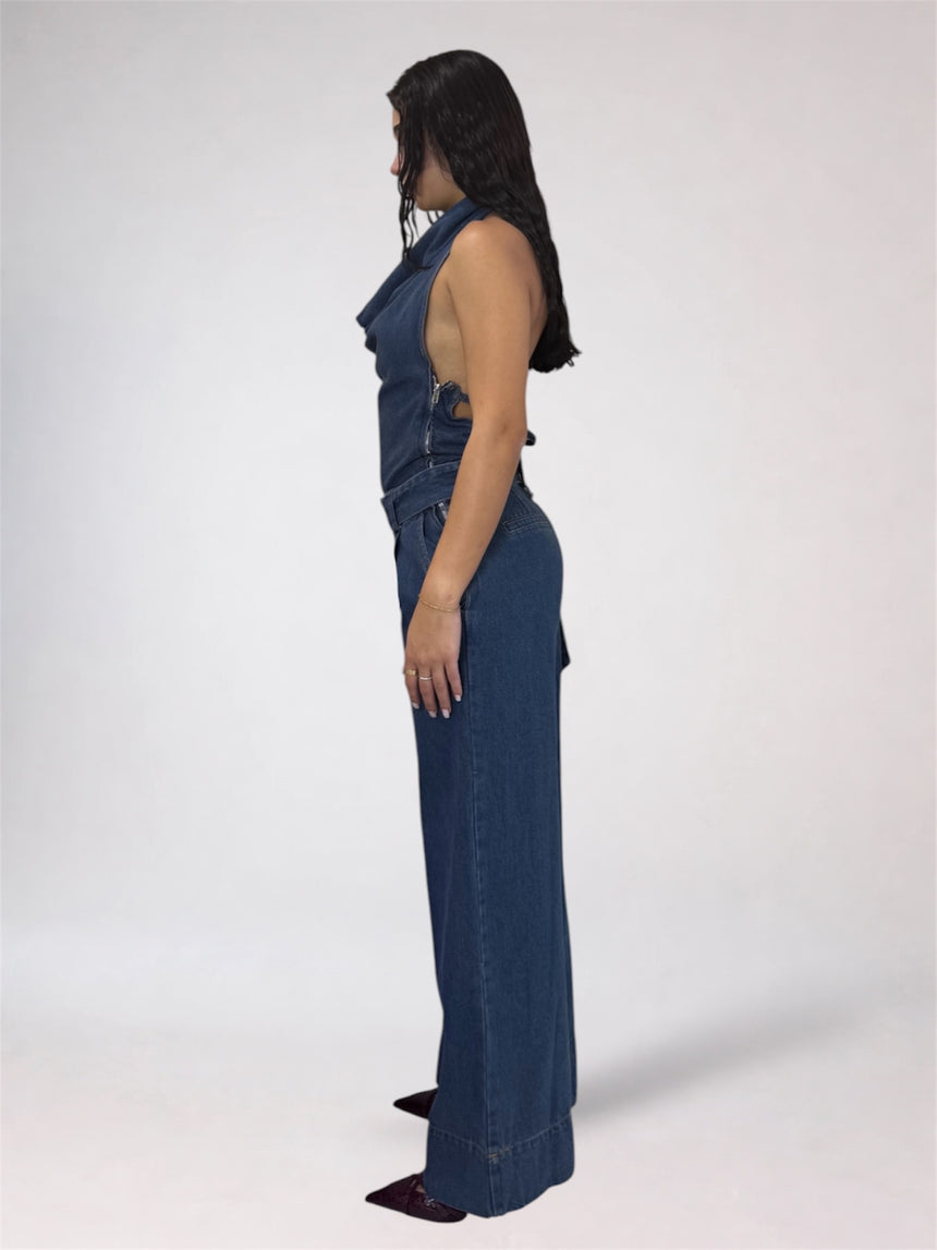 A woman stands in profile wearing a dark blue denim jumpsuit with a halter-style neckline and wide legs, photographed against a plain light gray studio background.