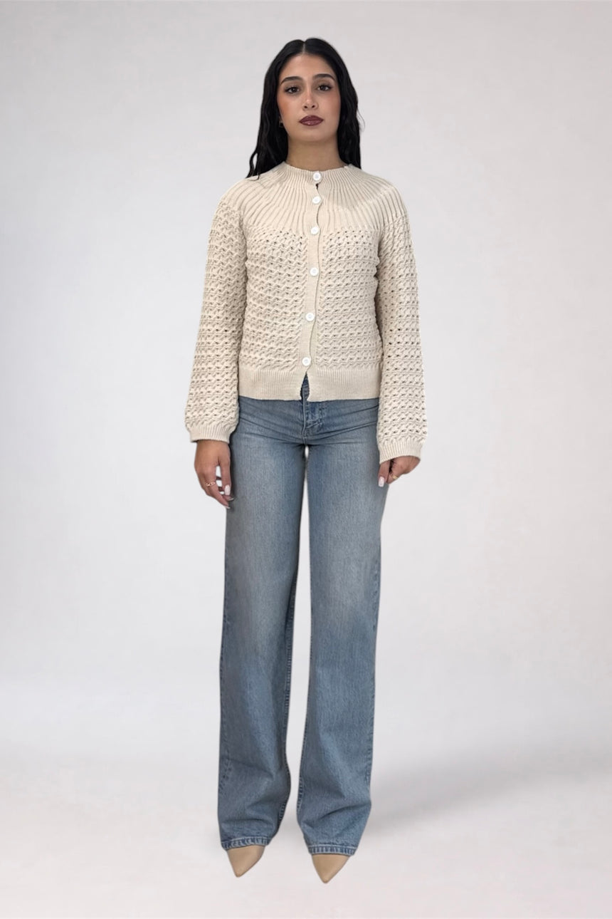 A woman stands against a light gray studio background wearing a cream-colored, long-sleeve knit cardigan with a button-up front and wide-leg blue jeans.