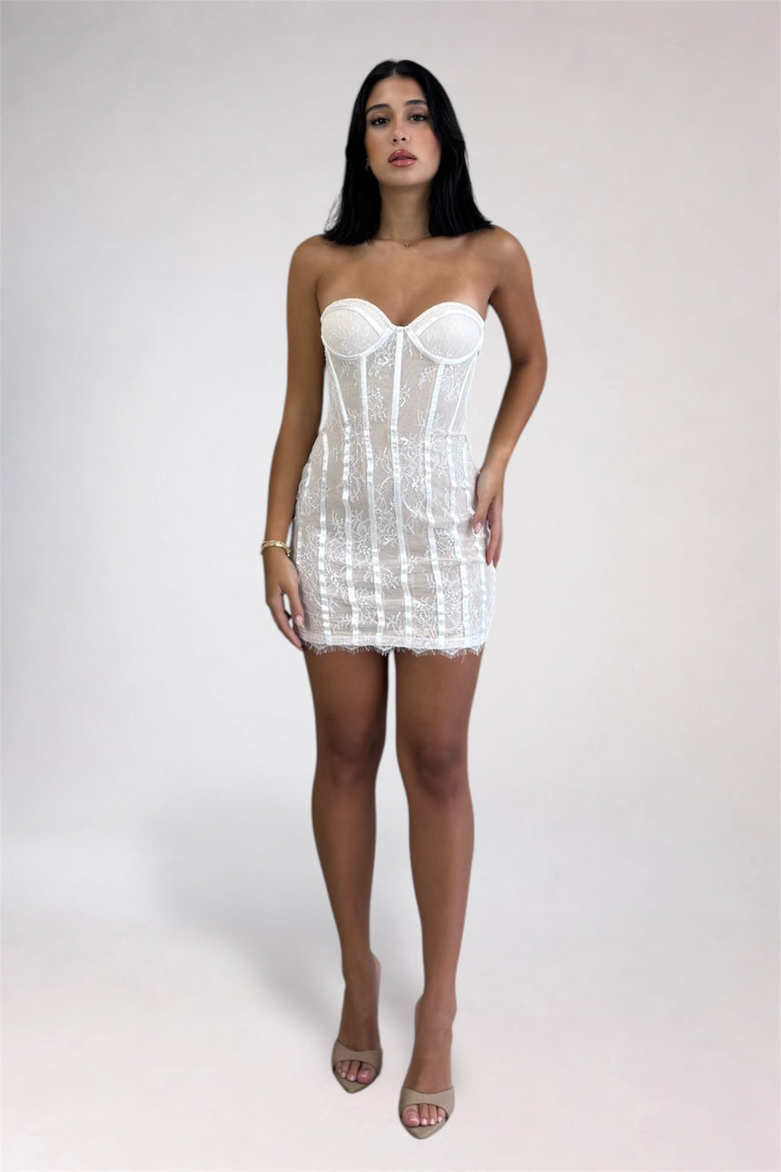 A woman modeling a white strapless corset mini dress with lace trim, standing against a light gray studio background.