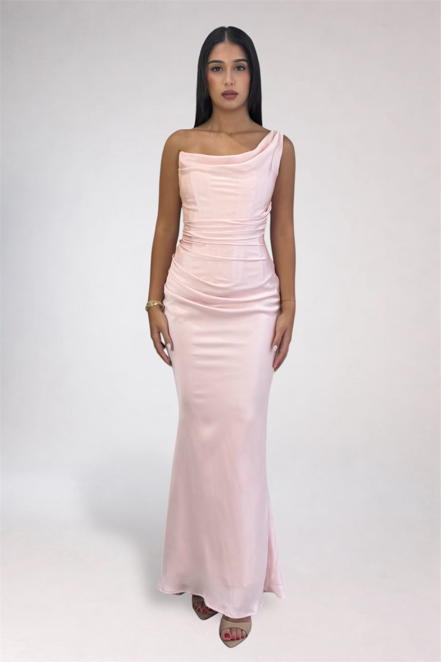 A woman modeling a light pink one-shoulder satin maxi dress against a plain light gray studio background.