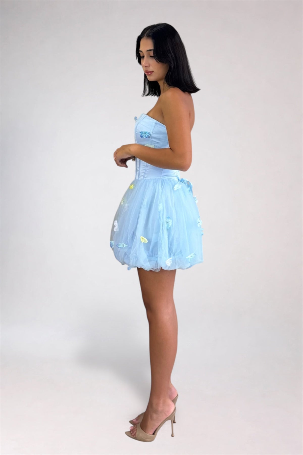 A woman modeling a light blue strapless tulle dress with a corset-style bodice and a flared skirt decorated with small white and yellow floral details, photographed against a plain light gray studio background.