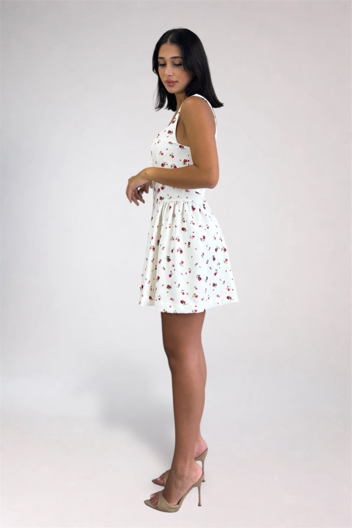 A woman modeling a sleeveless white mini dress with a small red and green cherry print, paired with nude high-heeled sandals, against a light gray studio background.