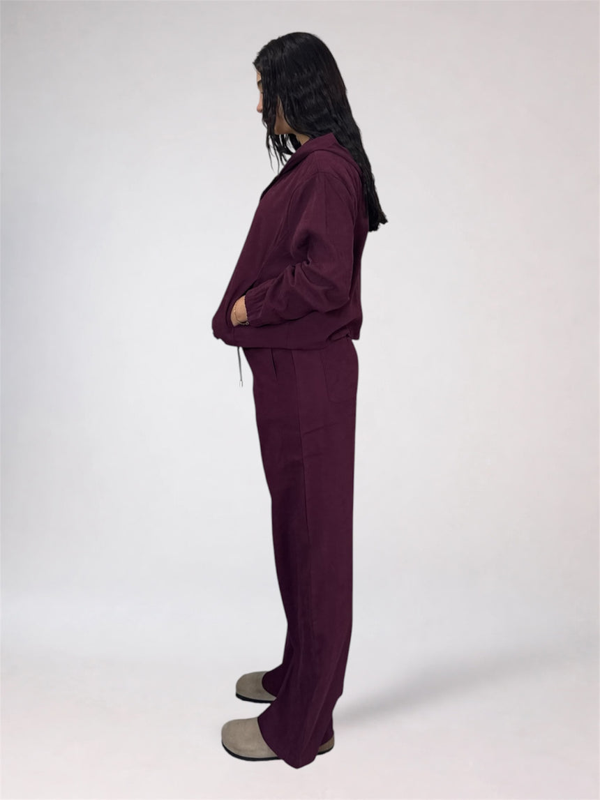 A model shown from the side profile wearing a matching deep plum purple lounge set consisting of a long-sleeve top and wide-leg pants, paired with gray slip-on shoes against a plain white studio background.