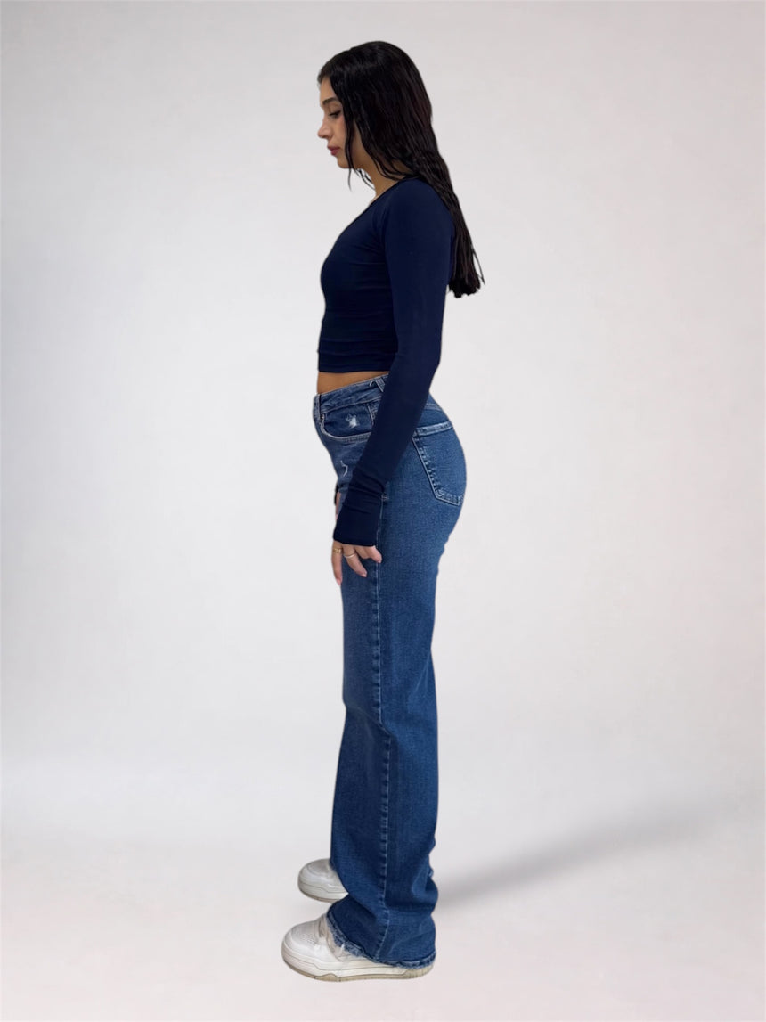 A woman shown in profile wearing a dark navy long-sleeve crop top, blue flare jeans, and white sneakers against a plain light gray studio background.