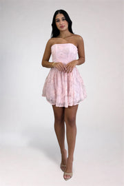 A woman modeling a strapless light pink lace mini dress with a flared skirt, paired with nude high-heeled sandals, against a plain light gray studio background.