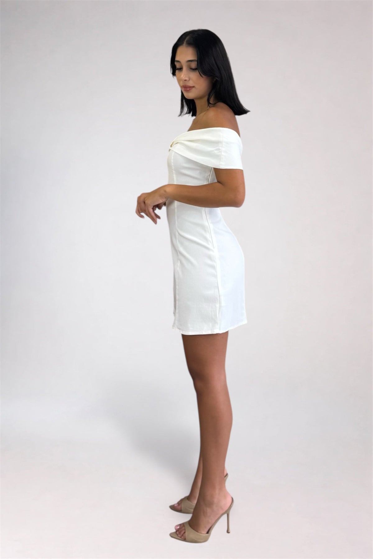 A woman modeling an off-the-shoulder white mini dress with a structured bodice and short skirt, paired with beige high-heeled sandals, against a light gray studio background.