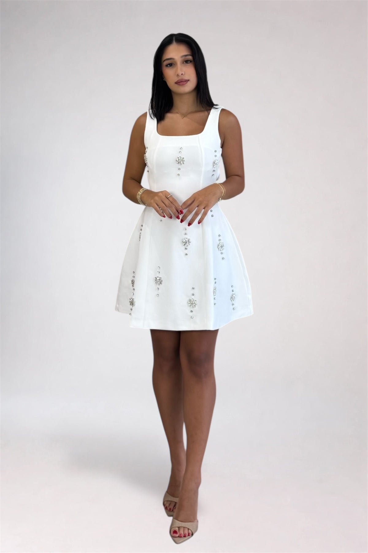 A woman modeling a white sleeveless mini dress with embroidered details, standing against a light gray studio background.