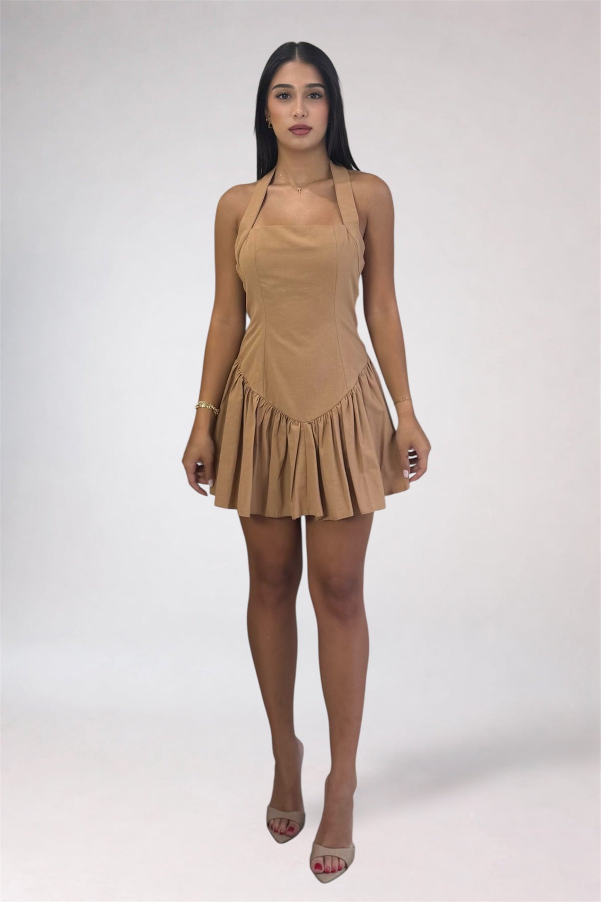 A woman modeling a tan halter-neck mini dress with a pleated ruffled skirt, paired with clear high-heeled sandals, against a light gray studio background.