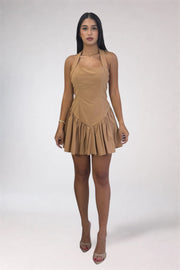 A woman modeling a tan halter-neck mini dress with a pleated ruffled skirt, paired with clear high-heeled sandals, against a light gray studio background.