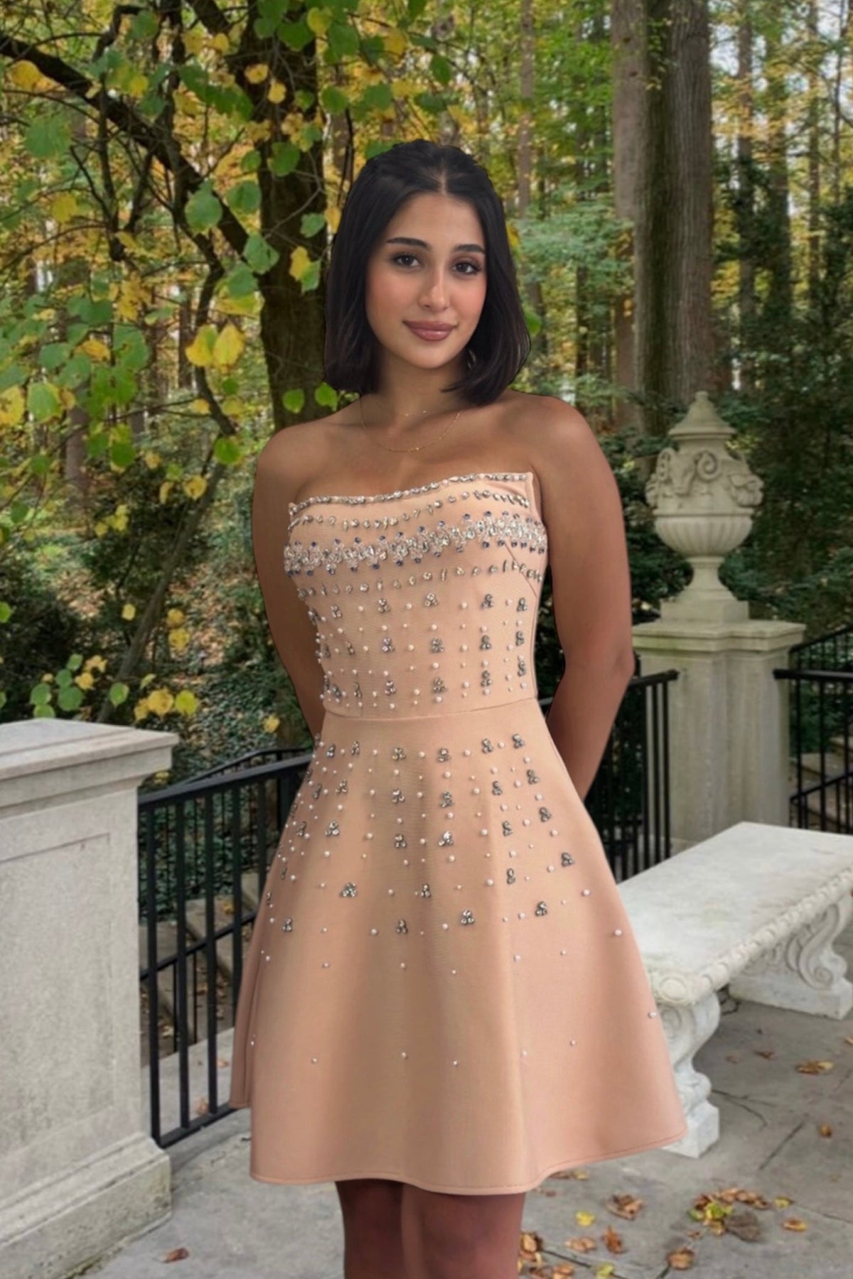 Woman in a beige strapless dress with embellishments standing outdoors in a garden setting.