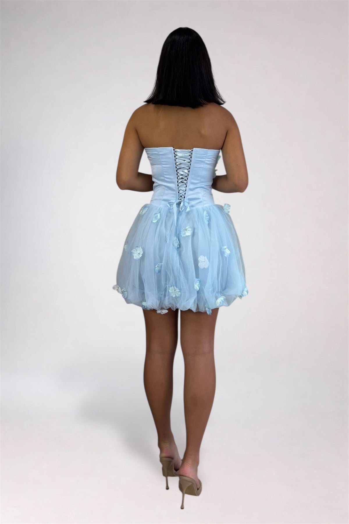 A woman viewed from behind wearing a strapless light blue tulle dress with a lace-up back and floral applique, paired with tan high heels, against a plain light gray studio background.