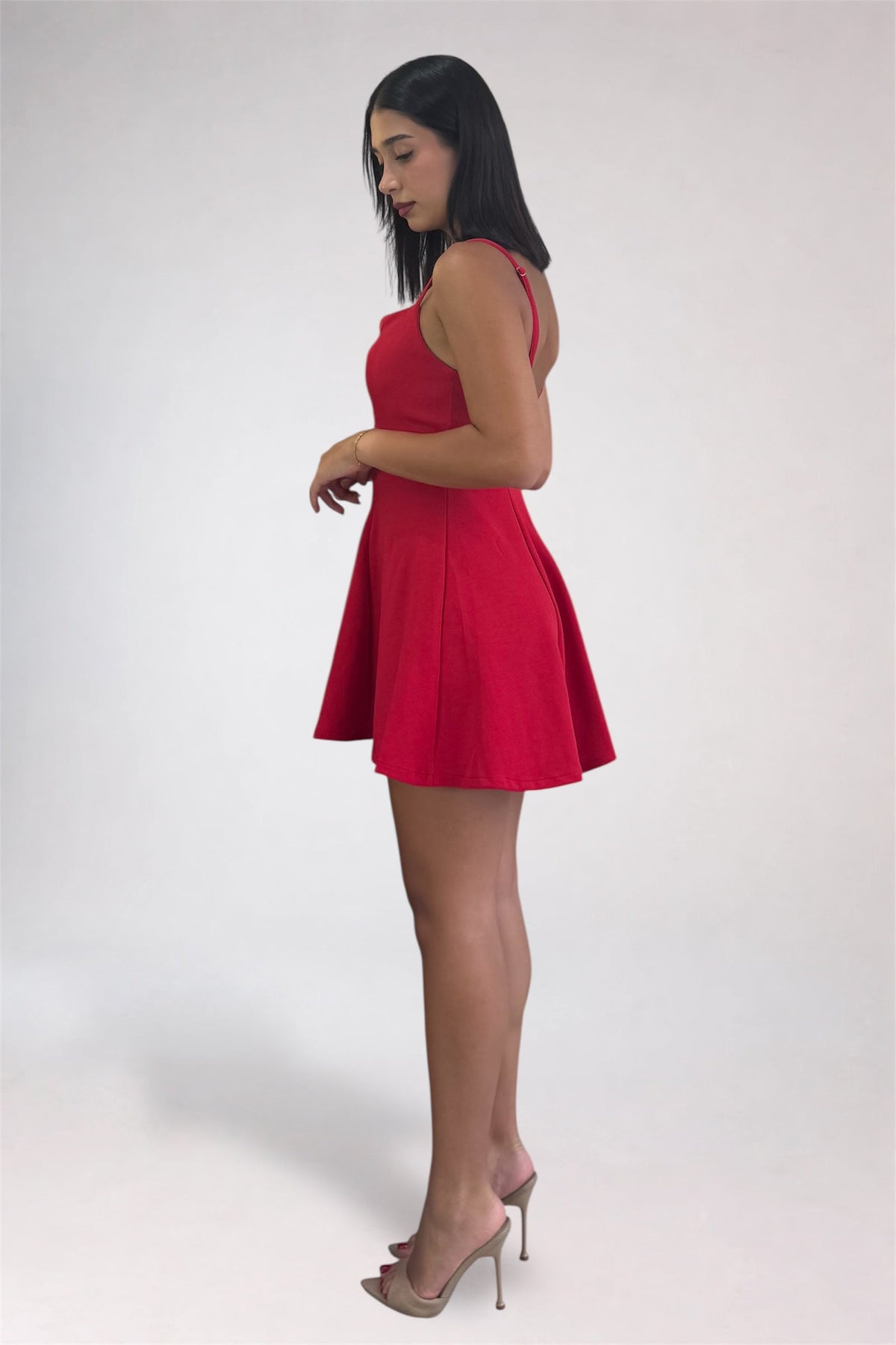 A woman modeling a bright red sleeveless mini dress with a flared skirt, paired with nude high-heeled sandals, standing against a plain light gray studio background.