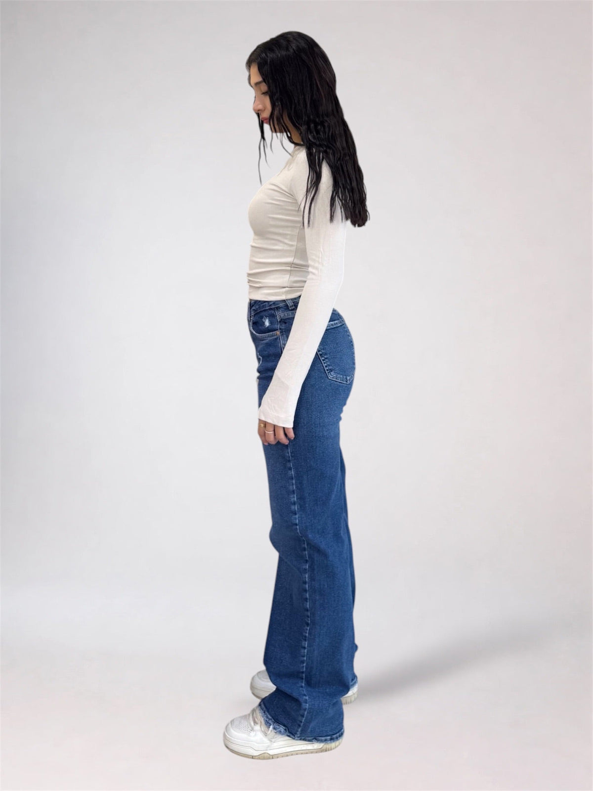A woman shown in profile wearing a fitted white long-sleeve top, blue wide-leg jeans, and white sneakers against a plain light gray studio background.