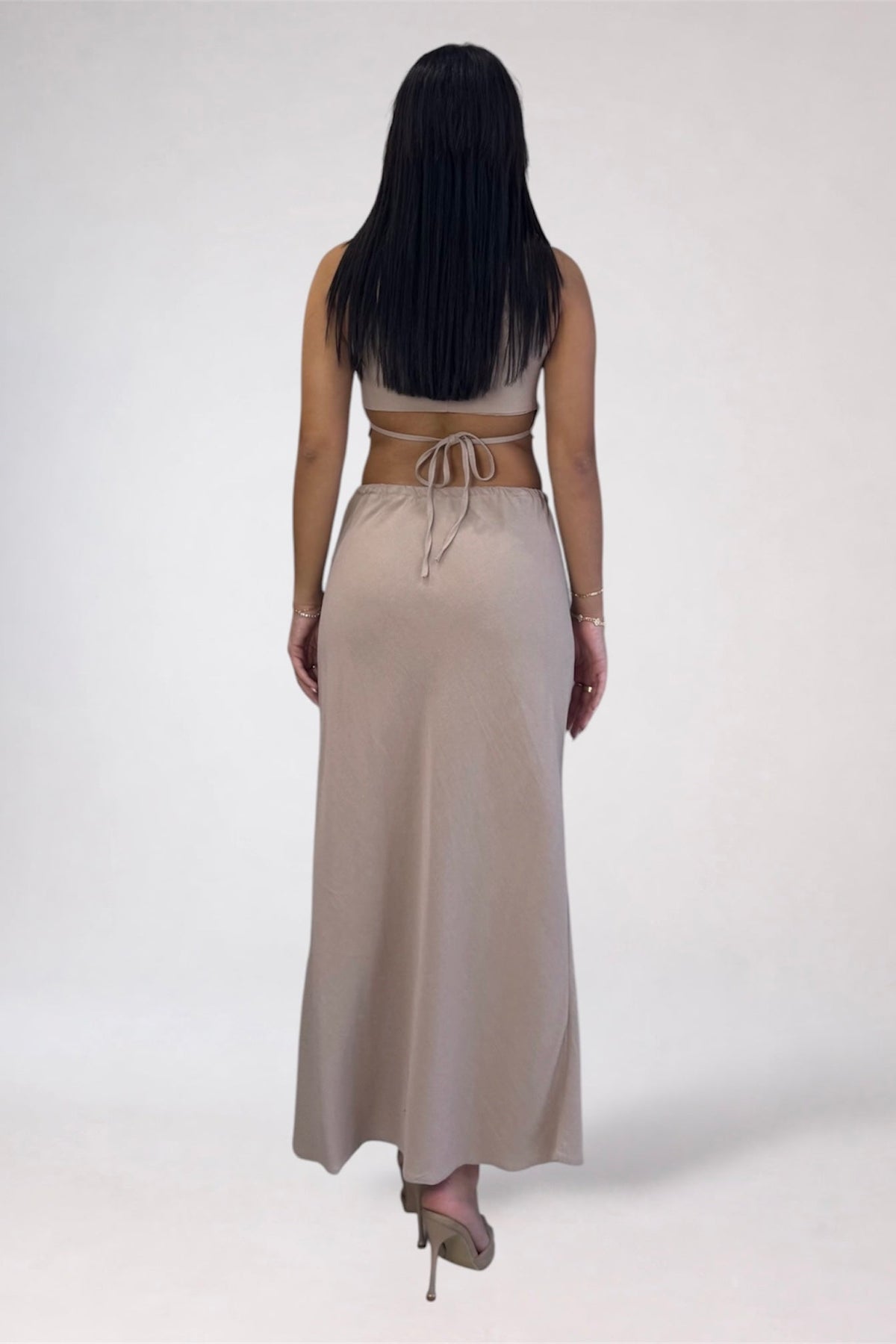 A woman viewed from behind wearing a beige two-piece set consisting of a sleeveless crop top with a tie waist and a matching maxi skirt, set against a plain light background.