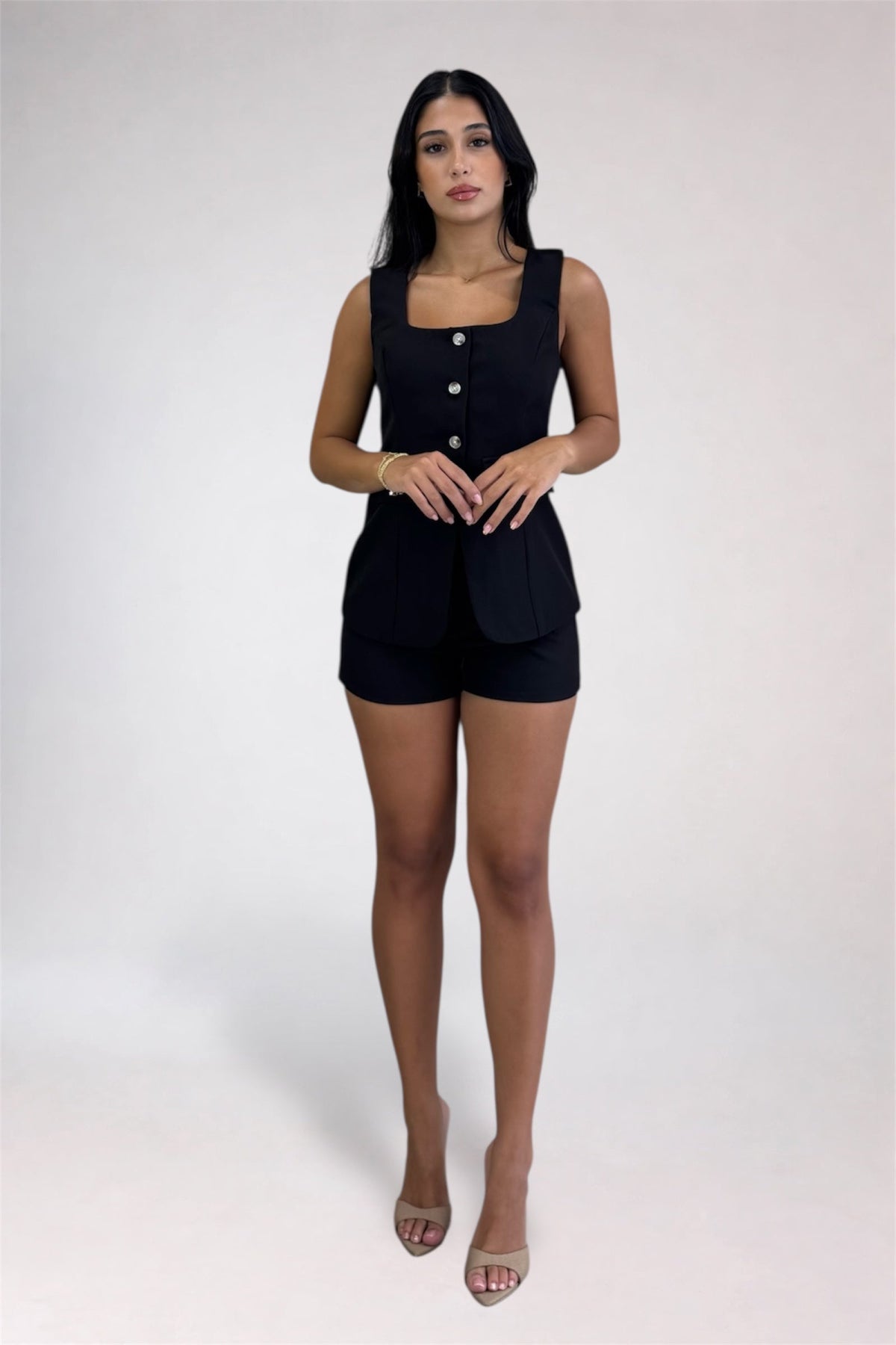 A woman modeling a black sleeveless romper with a square neckline, front button detail, and matching shorts, standing against a plain light gray studio background.
