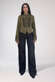 A model stands against a light gray studio background wearing a cropped olive green jacket with black lace-up detailing on the front and sleeves, paired with dark wide-leg jeans.