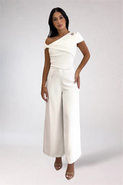 A woman modeling a white one-shoulder jumpsuit with wide-leg pants and nude high-heeled sandals against a light gray studio background.
