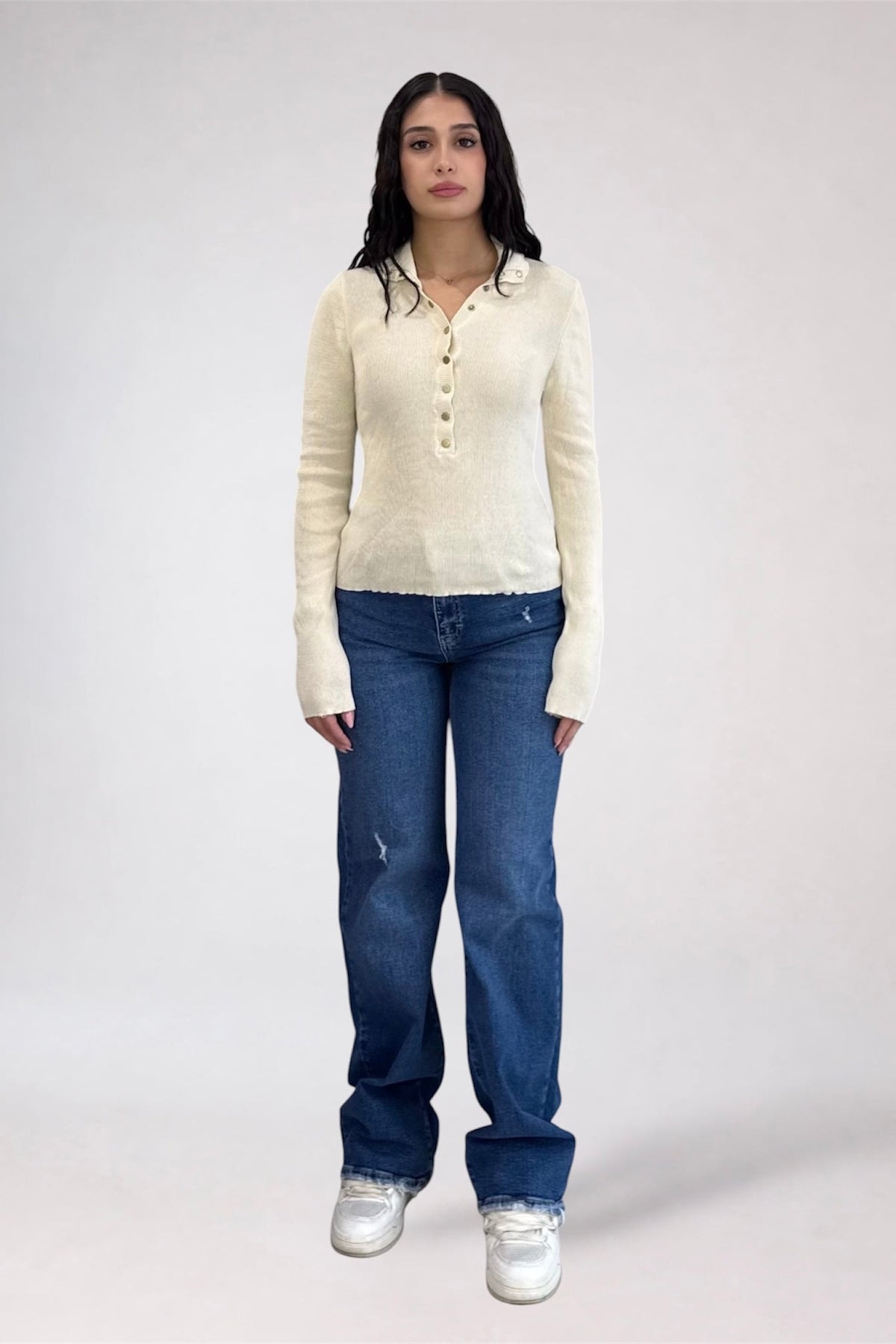 A woman stands against a light gray studio background wearing a cream ribbed knit long-sleeve henley top, blue jeans, and white sneakers.