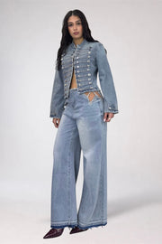 A model wearing a light blue denim set consisting of a cropped, embellished jacket with silver studs and matching wide-leg jeans, paired with dark pointed shoes, against a plain light gray studio background.
