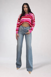 A woman modeling a pink and red striped cropped long-sleeve knit sweater paired with light-wash blue flare jeans, standing against a light gray studio background.